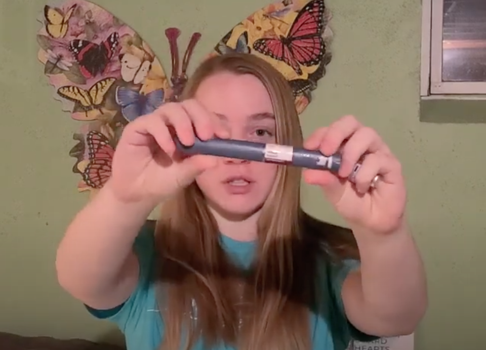 Teen girl with long brown hair wearing a blue shirt holds up blue insulin pen for camera.
