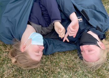 Picture shows two high school girls in their track sweats laying side by side on the ground and laughing.